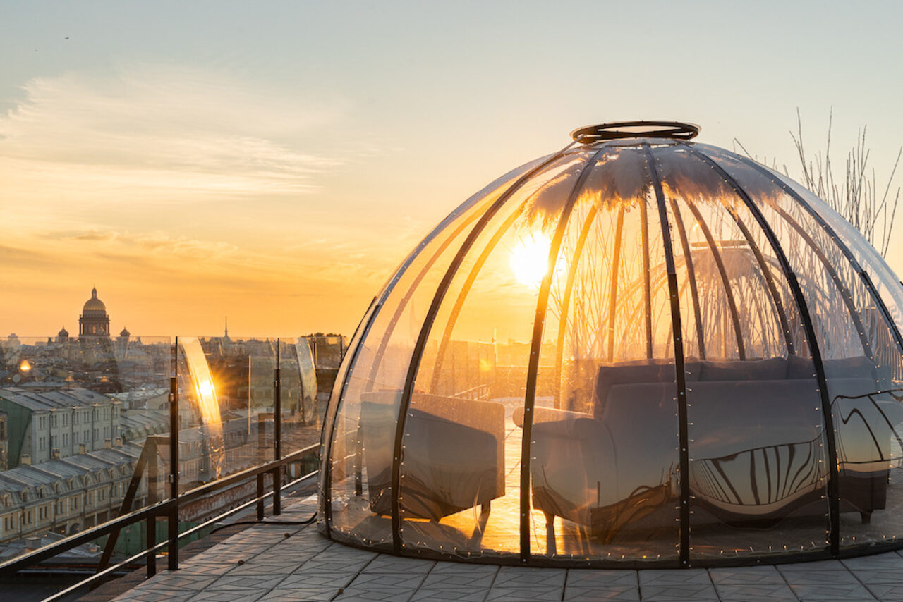 Арендовать Sky Terrasse(Небо Терраса). Купольная стеклянная конструкция на крыше с видом на город. Внутри находятся диваны. Прозрачные стены отражают солнечный свет. Пол выложен плиткой. Пространство окружено стеклянными ограждениями. Закатное солнце на горизонте.