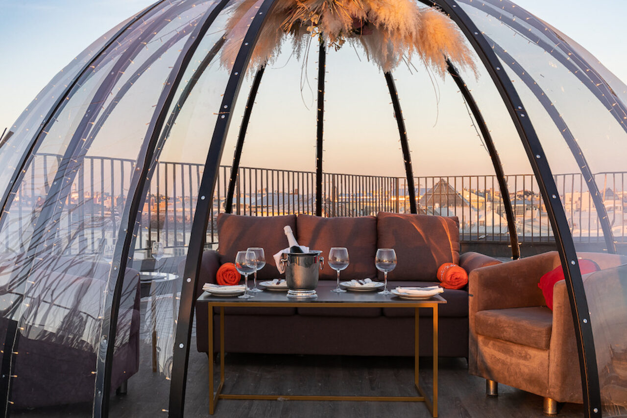 Арендовать Sky Terrasse(Небо Терраса). Современный купол из прозрачного материала на крыше, внутри коричневый диван и кресло, низкий стол с белыми тарелками, бокалами и ведёрком с бутылкой, украшение из сухих растений сверху, вокруг металлическое ограждение, вечернее освещение.
