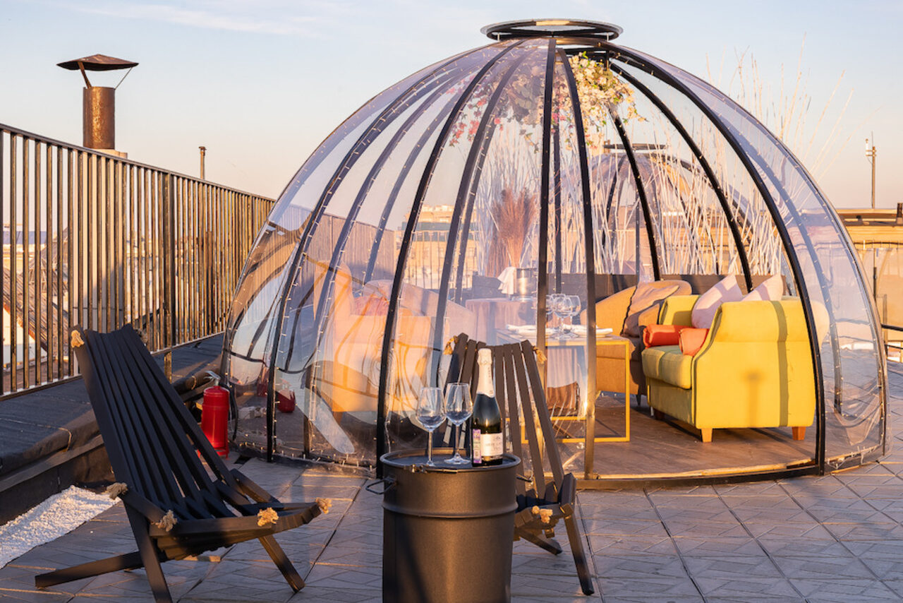 Арендовать Sky Terrasse(Небо Терраса). Купольная конструкция на террасе, прозрачные стены, внутри диваны и стол, на улице два черных кресла и столик с бутылкой и бокалами, рядом огнетушитель, растительность на заднем плане, металлическое ограждение, плиточное покрытие, теплое освещение.