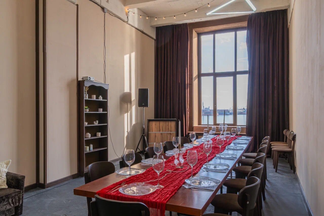 Арендовать Chocolate Loft (Шоколад Лофт). Зал с видом на Финский залив. Прямоугольное столовое пространство с длинным деревянным столом, накрытым красной скатертью и сервировкой на 12 персон. Металлические стулья, высокий стеллаж с посудой, светлые стены, большая штора, большое окно, черные портьеры, струнные светильники.