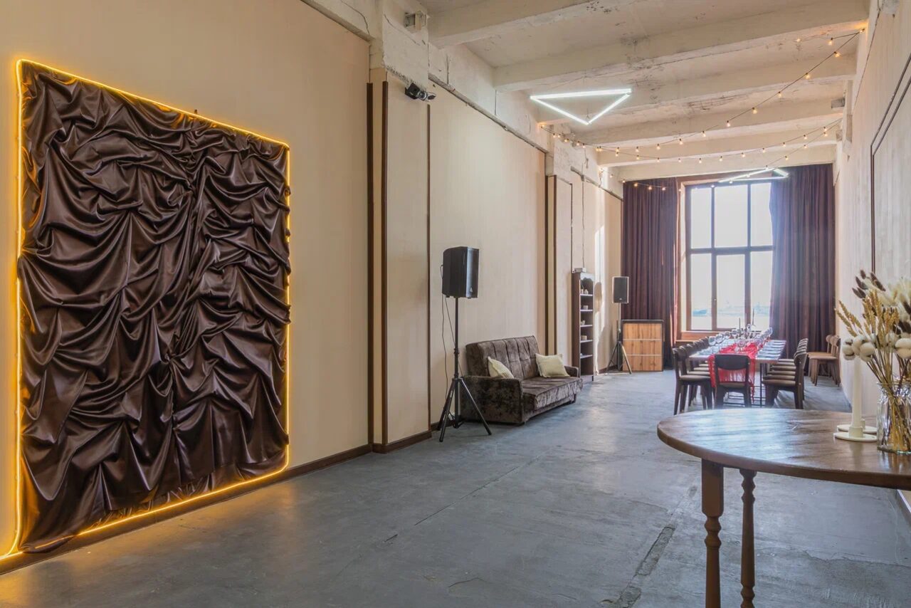 Арендовать Chocolate Loft (Шоколад Лофт). Зал с видом на Финский залив. Просторное помещение с высокими потолками, длинный стол с красным текстилем и стульями, диван, деревянный пол. Светлый потолок, стены с декоративной тканью в подсветке. Вертикальные окна с занавесками, треугольные светильники, дополнительные источники света на потолке.