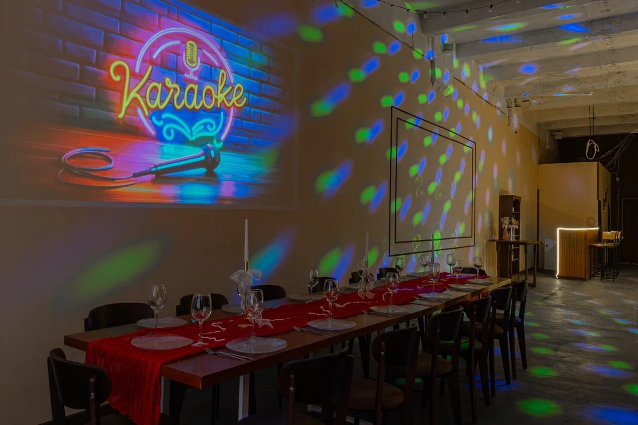 Арендовать Chocolate Loft (Шоколад Лофт). Зал с видом на Финский залив. Просторное помещение с длинным деревянным столом, покрытым красной дорожкой, сервировано бокалами и свечами. На стене неоновая надпись "Karaoke" и микрофон. Тёмные стулья, разноцветное освещение, белый кирпич, барная стойка в глубине.
