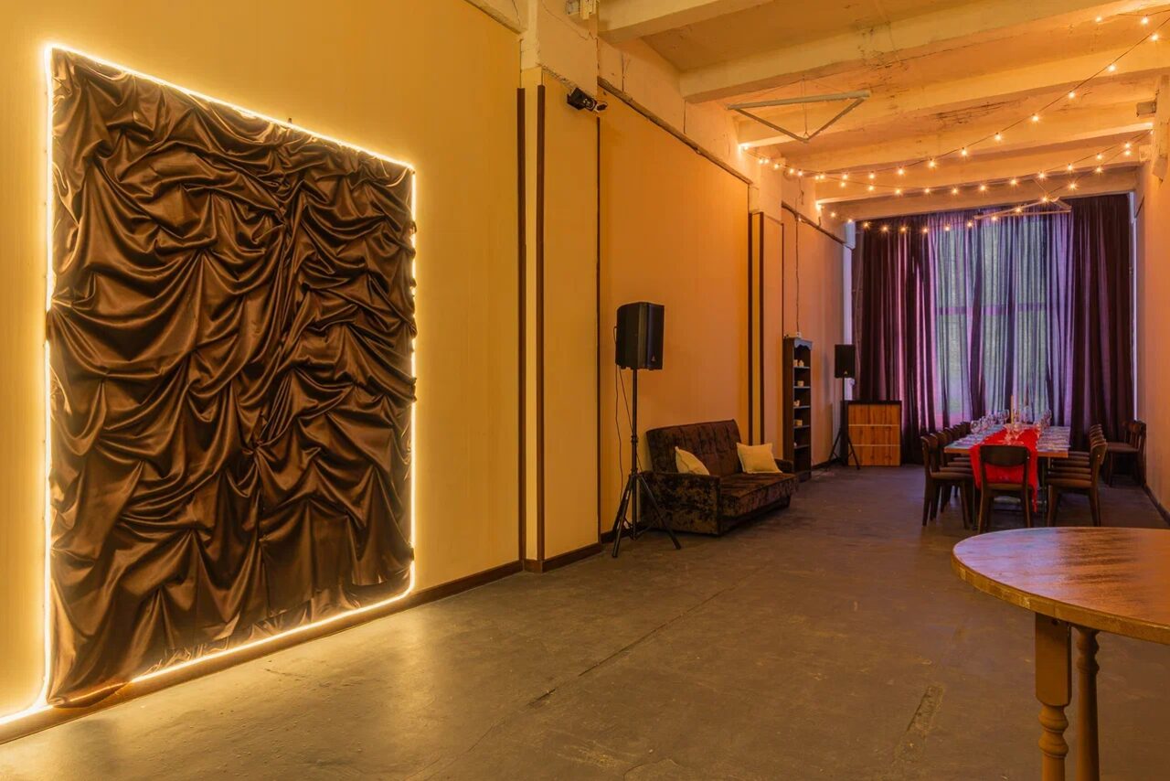 Арендовать Chocolate Loft (Шоколад Лофт). Зал с видом на Финский залив. Лофт-пространство с высоким потолком, обеденным столом и стульями. Тёмные шторы на окнах, драпировка на стене с подсветкой, диван, акустические колонки. Потолочное освещение из гирлянд, пол тёмно-серый, стены бежевые.