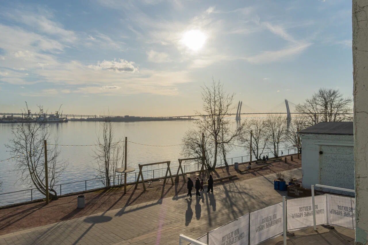 Арендовать Chocolate Loft (Шоколад Лофт). Зал с видом на Финский залив. Променад вдоль реки с асфальтированной дорожкой, несколько деревьев без листвы, лавочки, находящиеся на солнце, вдалеке мост, спокойная вода, небо с облаками, группа людей, ограждение с надписями, металлические конструкции.
