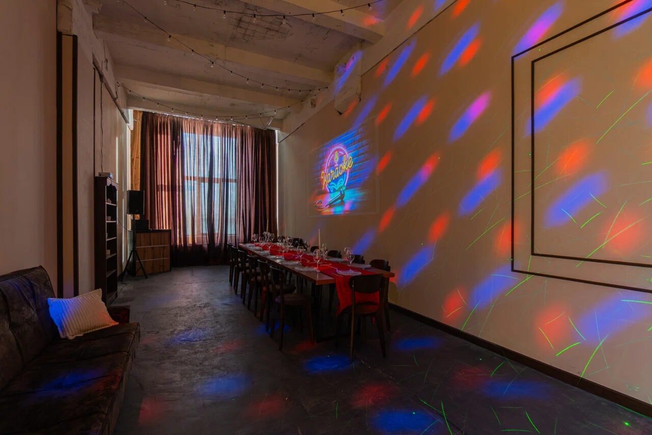 Арендовать Chocolate Loft (Шоколад Лофт). Зал с видом на Финский залив. Пространство с длинным столом и стульями, оформленное для банкета, стены украшены световыми проекциями с красными и синими узорами, тёмный пол, диван сбоку, большие окна с занавесками. На стене проекция круглой формы с текстом.