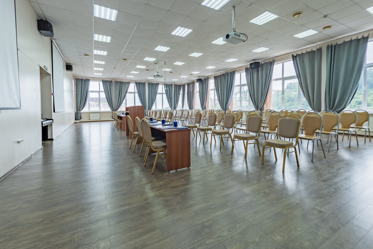 Арендовать Конференц зал в отеле Гарден Хиллс 3. Большая комната с деревянным полом, кафельным потолком с встраиваемыми светильниками, длинными рядами стульев и деревянными столами. Большие окна с серыми шторами, проекторы, экраны и кондиционеры на стенах.