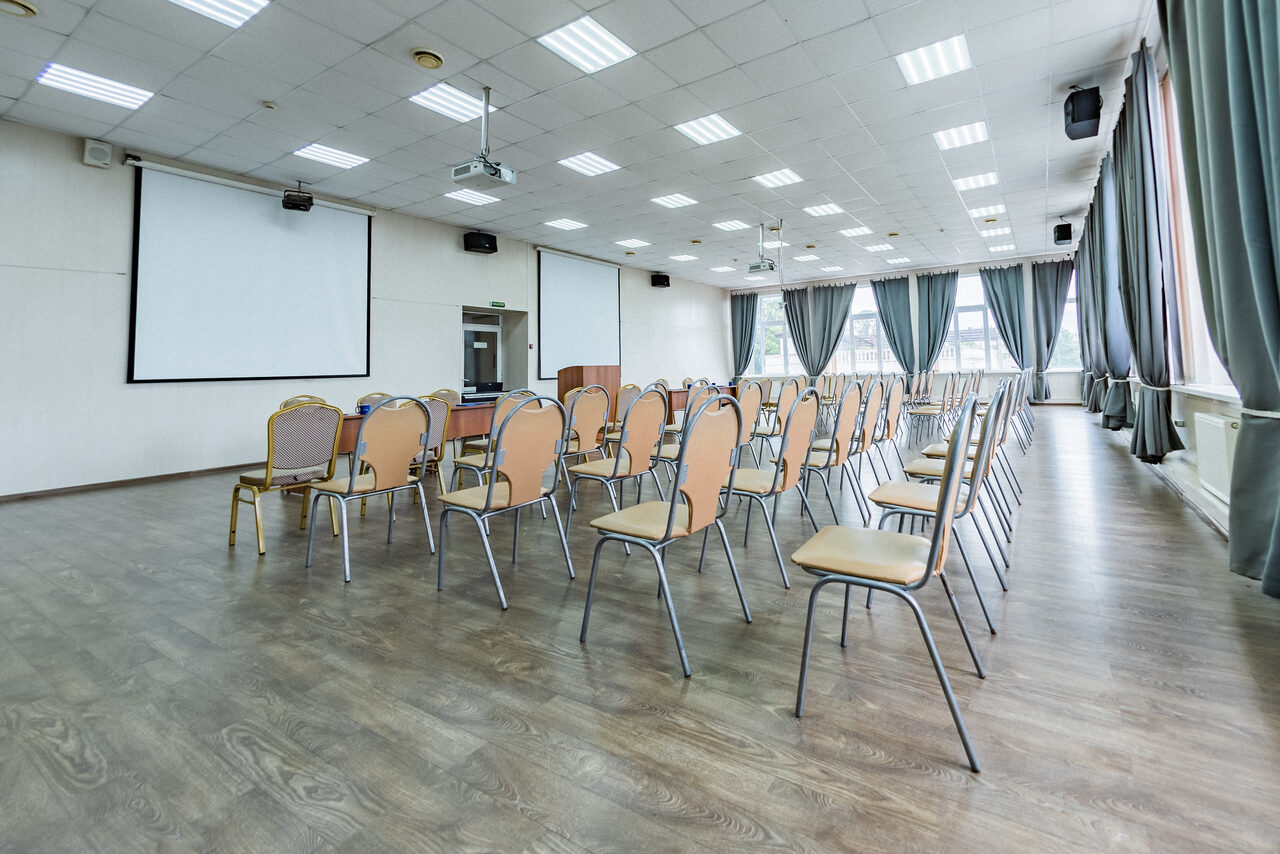 Арендовать Конференц зал в отеле Гарден Хиллс 3. Конференц-зал с деревянным полом, белыми стенами и подвесным потолком. Ряды стульев, проекционный экран, проекторы, аудиоколонки, большие окна с серыми шторами. Посредине зала установлена кафедра.