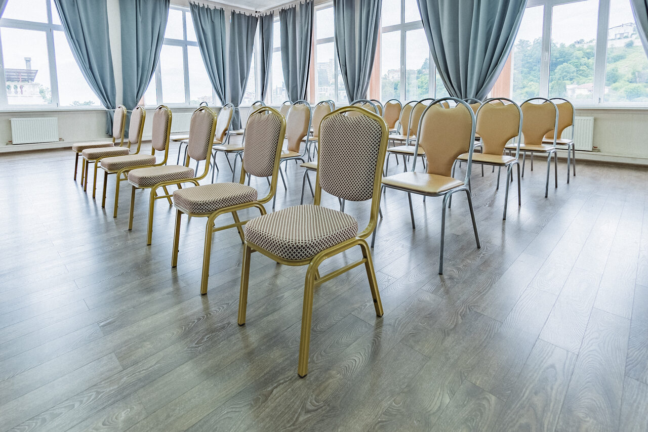 Арендовать Конференц зал в отеле Гарден Хиллс 3. Просторное помещение с рядами стульев бежевого цвета. Пол из светлого дерева, большие окна с серыми шторами. Светлые стены, естественное освещение из окон. Стулья расположены в несколько рядов.