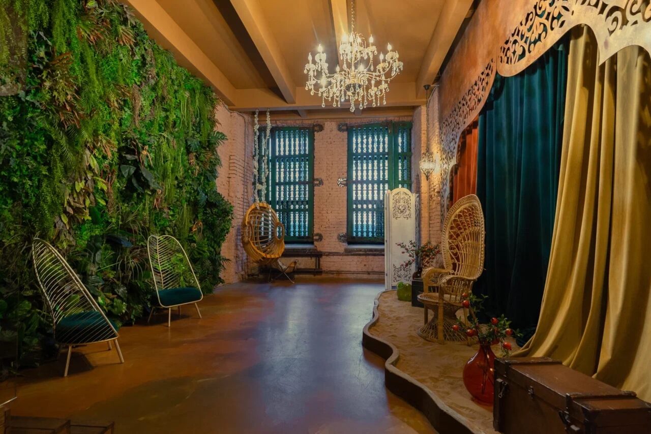 Арендовать Polygon(Полигон). Зал Марокко. Пространство с зелёной стеной из растений, двумя креслами-коконами, качелями, сценой с драпировкой и деревянным декором. Потолок с открытыми балками, люстра и настенные светильники. Цвета: зелёный, бежевый, коричневый. Пол бетонный, стены из кирпича.