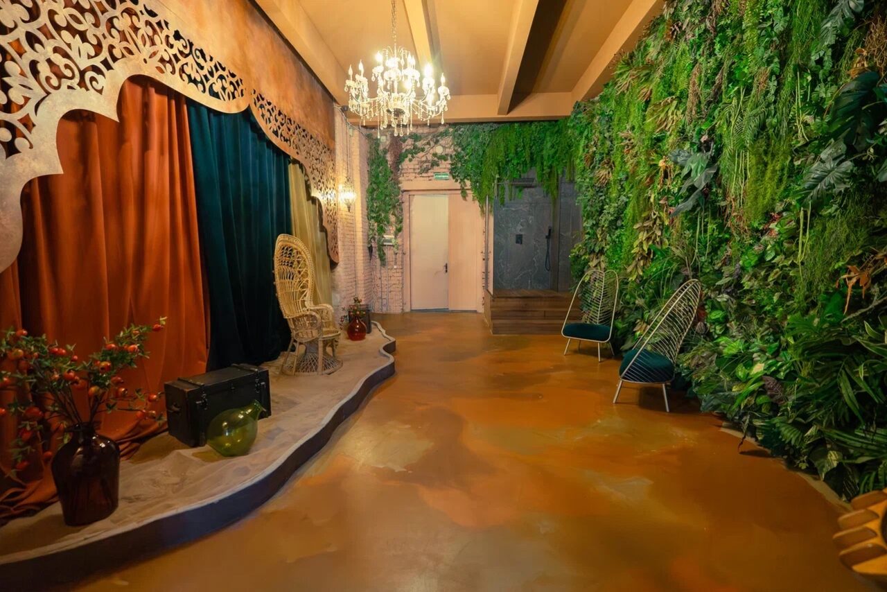 Арендовать Polygon(Полигон). Зал Марокко. Пространство с деревянным полом коричневого цвета, на стенах зелёные растения. Справа два стула с металлическими спинками, слева плетёное кресло, драпировки оранжевого и тёмно-зелёного цвета. Декоративная люстра, деревянный декор на стене.