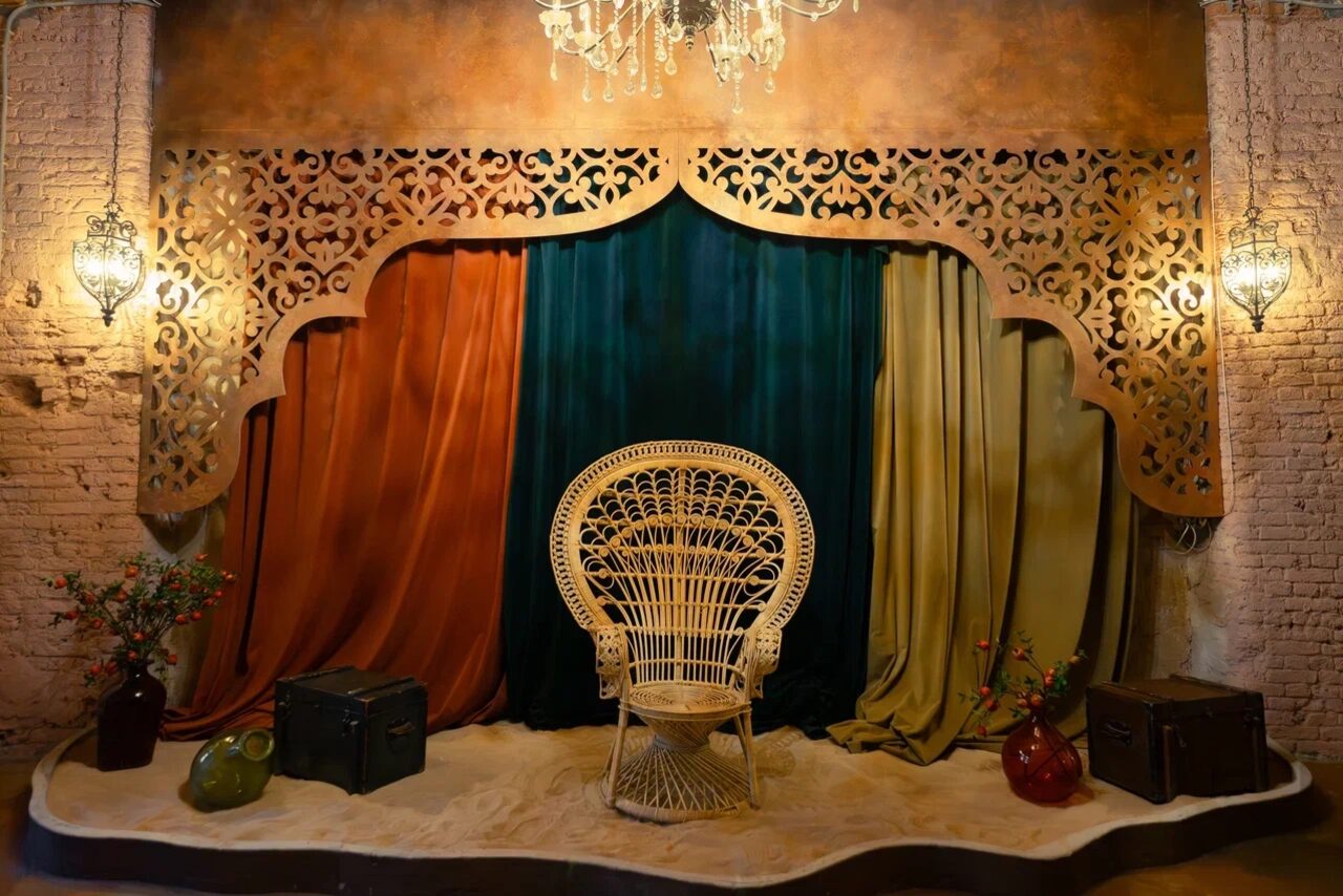 Арендовать Polygon(Полигон). Зал Марокко. Декорированное пространство с узорной аркой, драпированное тканями оранжевого, синего и зелёного цветов. Плетёное кресло в центре. По бокам коричневые чемоданы и вазы с цветами. Стены из светлого кирпича. Подвесные фонари и декоративная люстра.