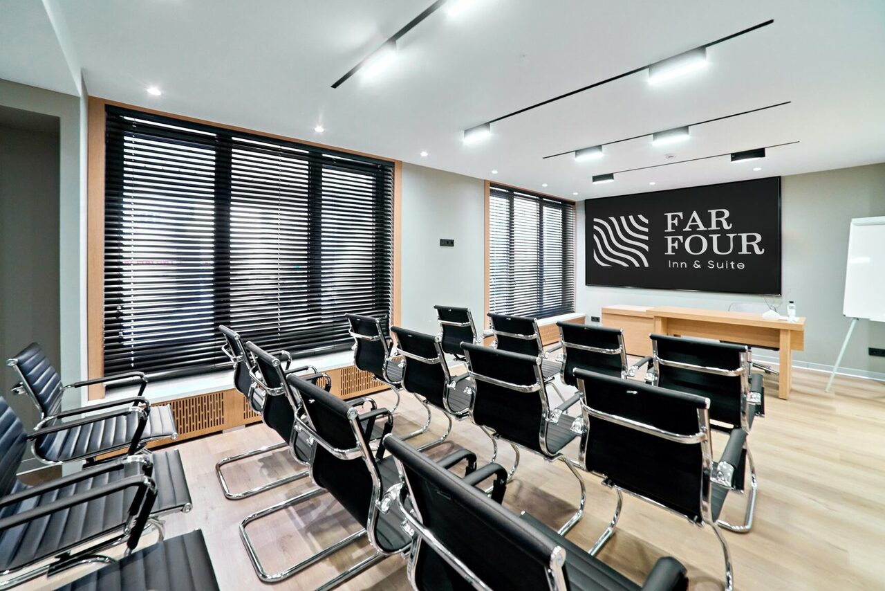 Арендовать FarFour (ФарФор). Конференц-зал. Конференц-зал с рядами черных кресел на металлокаркасе, светлые деревянные полы, белая доска, деревянный стол на подиуме, окна с жалюзи, встроенное потолочное освещение, на стене надпись «FAR FOUR inn & Suite».