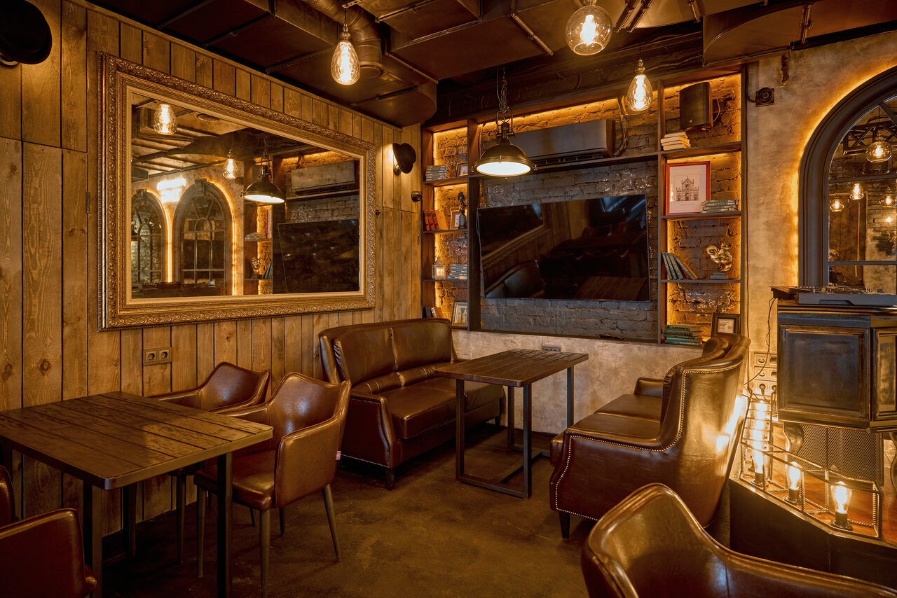 Арендовать Tangiers Lounge(Танжер Лаундж) . Уютное помещение с деревянными стенами, коричневыми кожаными креслами и диванами, столами из темного дерева. На стенах — зеркала и полки с книгами. Подвесное освещение с лампами накаливания, арочные окна. Атмосферный полумрак.