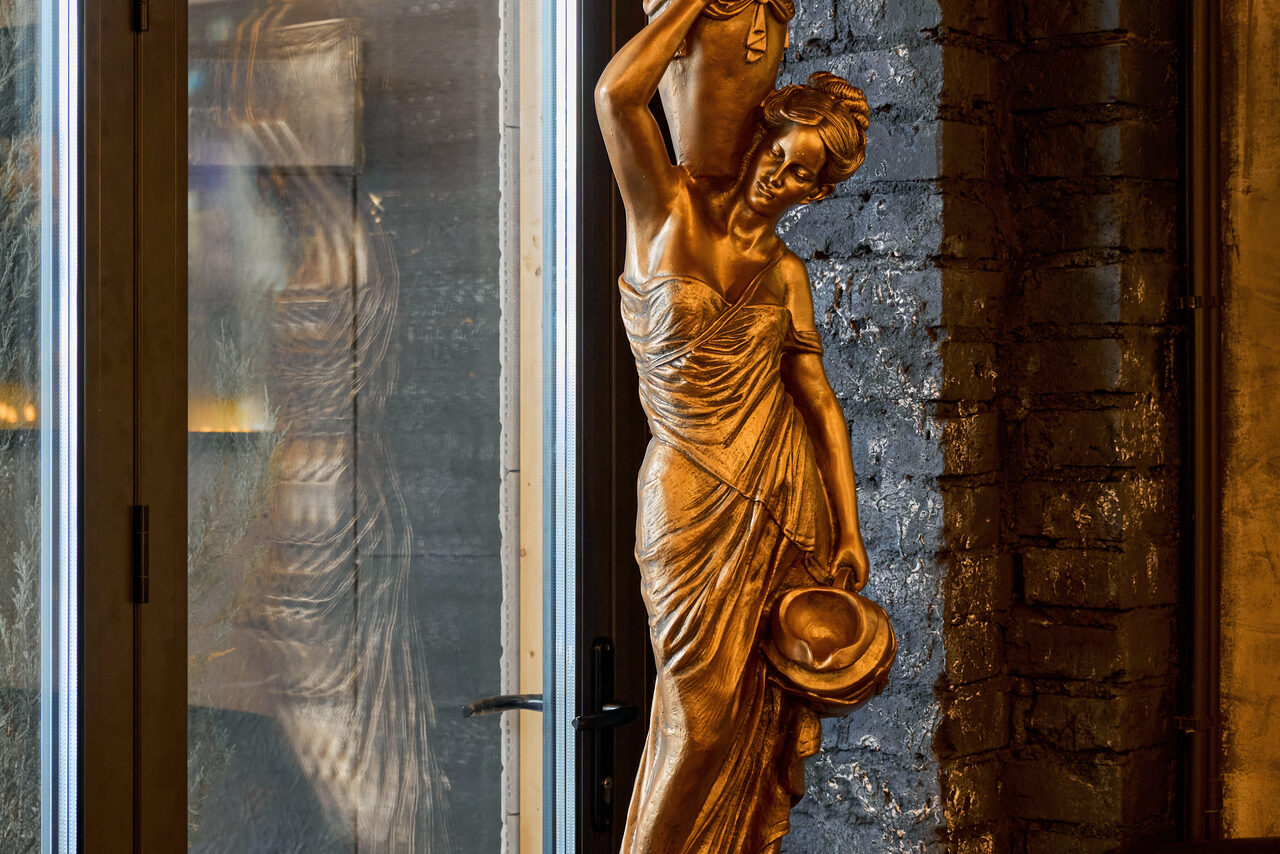 Арендовать Tangiers Lounge(Танжер Лаундж) . Статуя женщины из бронзового материала рядом с кирпичной стеной и стеклянной дверью. Женщина держит кувшин над головой и шляпу в другой руке. Теплое освещение.