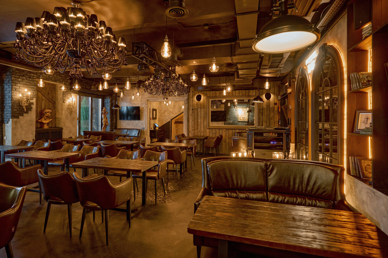 Арендовать Tangiers Lounge(Танжер Лаундж) . Зал с деревянными столами и кожаными стульями. Потолок украшен люстрами и лампами с теплым освещением. Стены декорированы кирпичом и деревом, есть большие оконные проемы. На стенах полки с книгами и картинами. Теплые коричневые и бежевые тона.