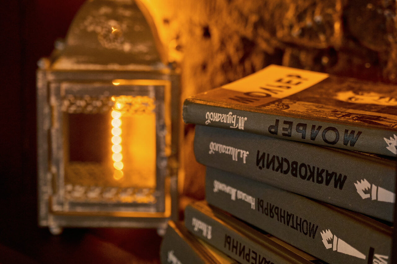 Арендовать Tangiers Lounge(Танжер Лаундж) . Книжная полка с книгами на русском языке в твердом переплете, рядом металлический фонарь. Теплое освещение, текстурная стена из камня. Книги сложены стопкой. Цветовая гамма в коричневых и золотистых оттенках.