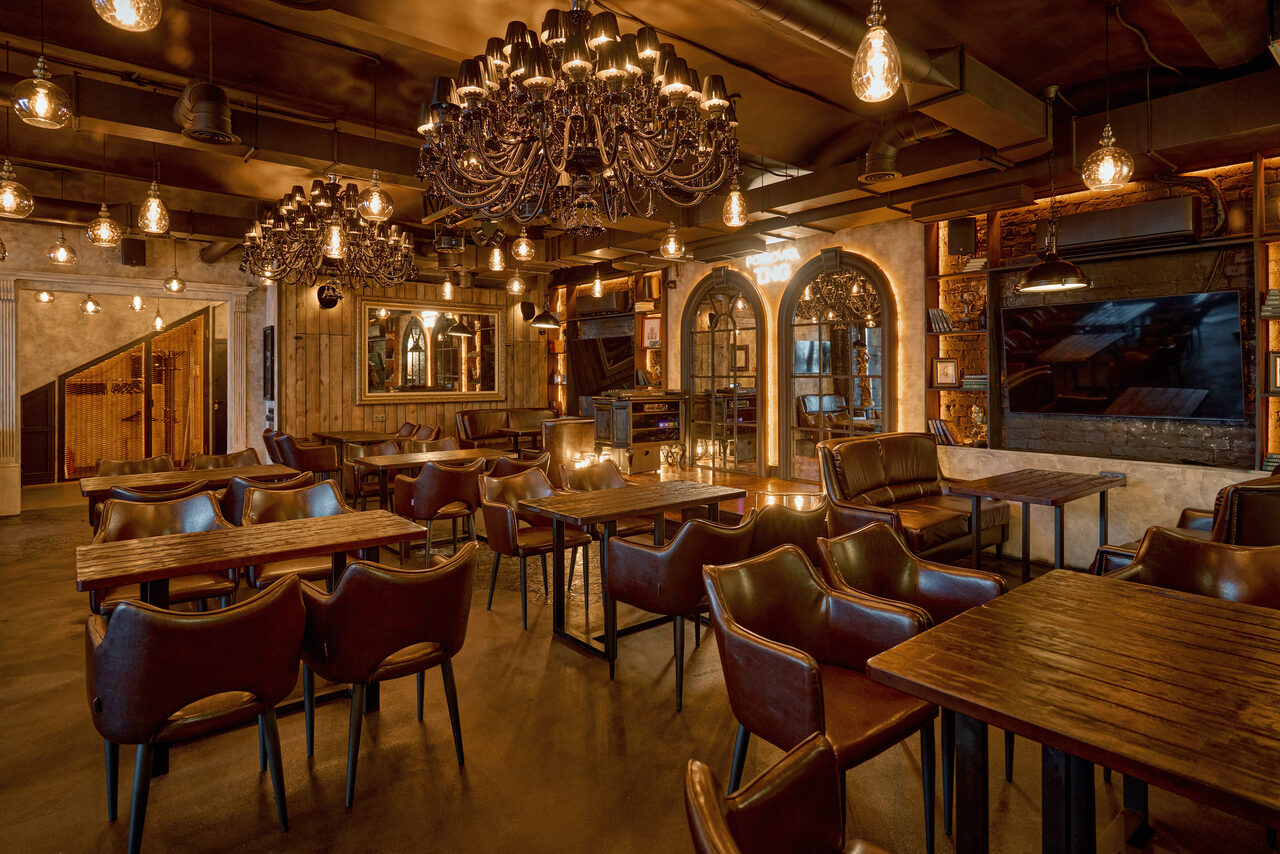 Арендовать Tangiers Lounge(Танжер Лаундж) . Ресторанное пространство с деревянными столами и кожаными стульями, винтажными люстрами и индустриальными светильниками. На стенах зеркала и декоративные арки. Темные стены и полы, уютное освещение, комплексная отделка в стиле лофт.