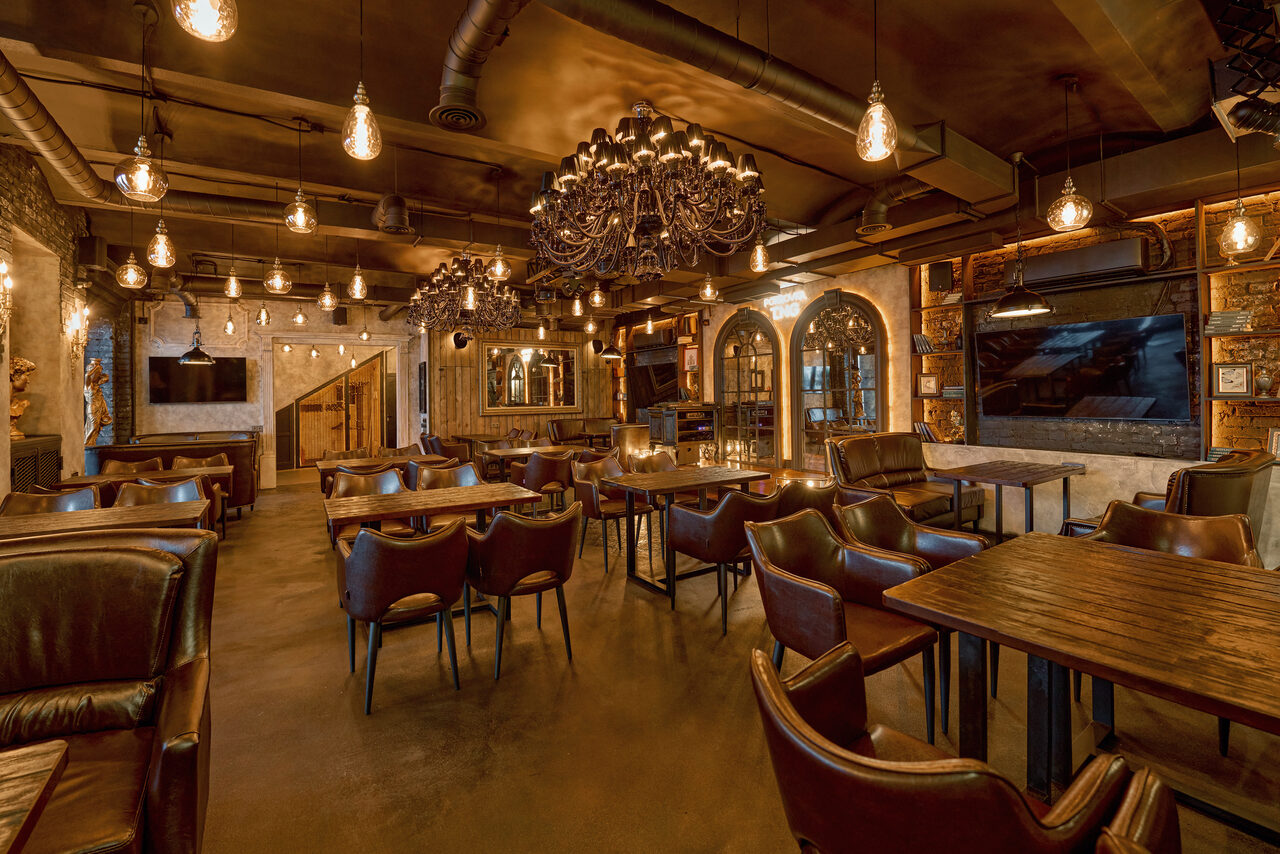 Арендовать Tangiers Lounge(Танжер Лаундж) . Кафе с деревянными столами и кожаными стульями, подвесные лампы и большая люстра, темные стены и пол теплыми тонами, арочные окна, телевизор на стене, кирпичные акценты, настроенное освещение, декор в индустриальном стиле.