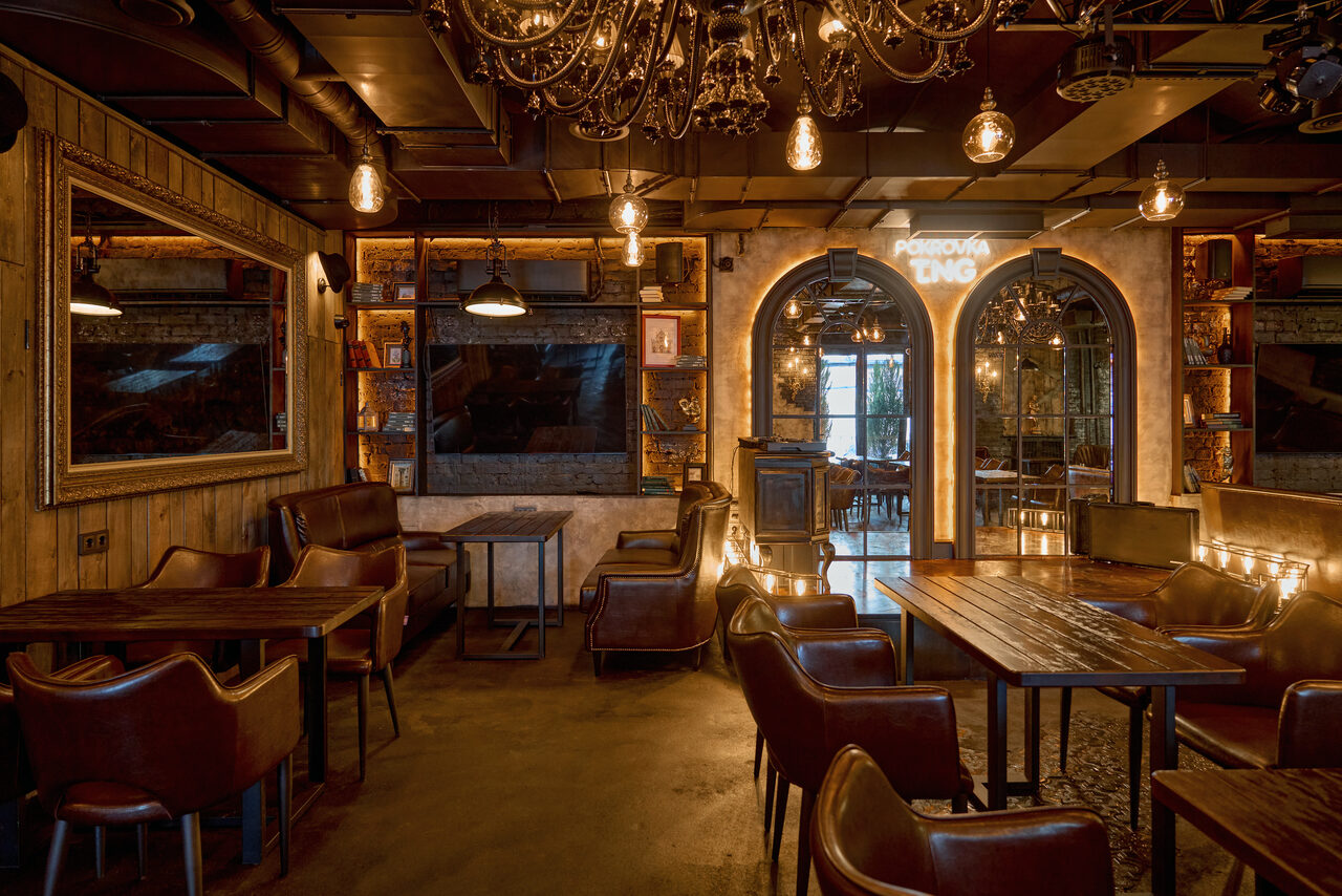 Арендовать Tangiers Lounge(Танжер Лаундж) . Уютное кафе с деревянными столами и кожаными стульями, освещение в виде множества ламп, стены украшены зеркалами и деревянными панелями, арочные двери, пол бетонный, темный цветовой тон, на одной из арок светящаяся надпись.