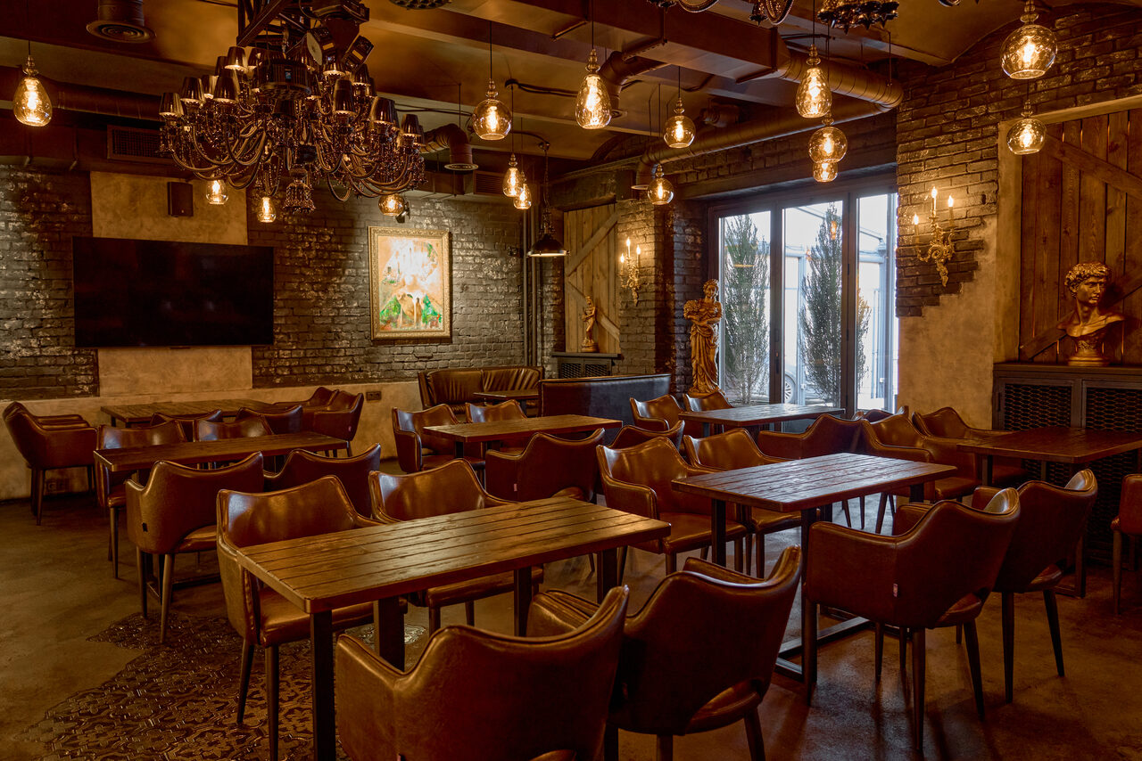 Арендовать Tangiers Lounge(Танжер Лаундж) . Пространство в стиле кафе с деревянными столами и коричневыми кожаными креслами, темные кирпичные стены, большой экран, картины, стеклянная дверь с видом на улицу. Потолок украшен черными люстрами и лампами накаливания.