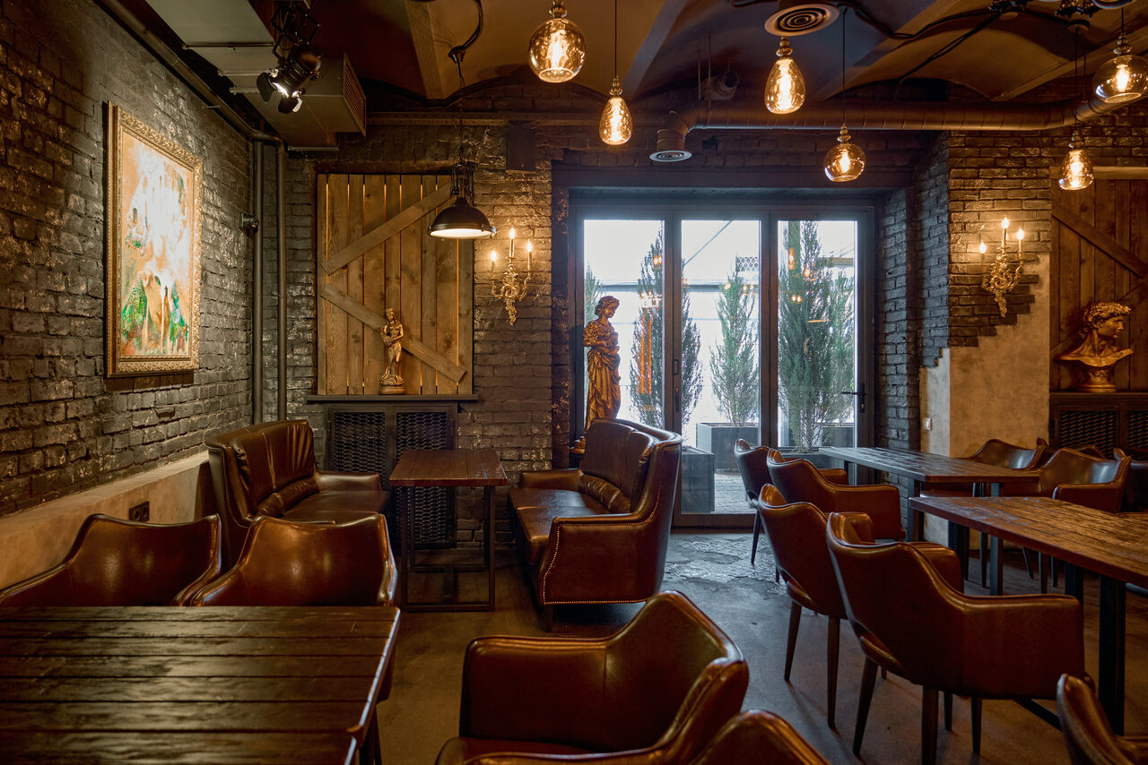 Арендовать Tangiers Lounge(Танжер Лаундж) . Интерьер с кирпичными стенами, темными деревянными столами и коричневыми кожаными креслами. Потолок с подвесными лампами, большие окна с дверью. На стенах картины и скульптуры, деревянная панель в стиле амбарной двери. Эффектная атмосфера с теплым освещением.