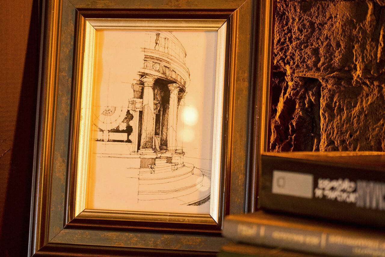 Арендовать Tangiers Lounge(Танжер Лаундж) . Картина с архитектурным эскизом в золоченой рамке, рядом стопка книг. Стена из каменной кладки с теплым освещением. Оттенки коричневого и золотого.