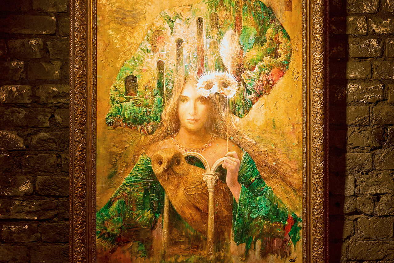 Арендовать Tangiers Lounge(Танжер Лаундж) . Картина в декоративной рамке на фоне кирпичной стены. На картине изображена женщина с совой, окруженная зелеными и золотистыми элементами, держащая цветы. Тон освещения теплый, рамка резная, кирпичи темно-коричневые.