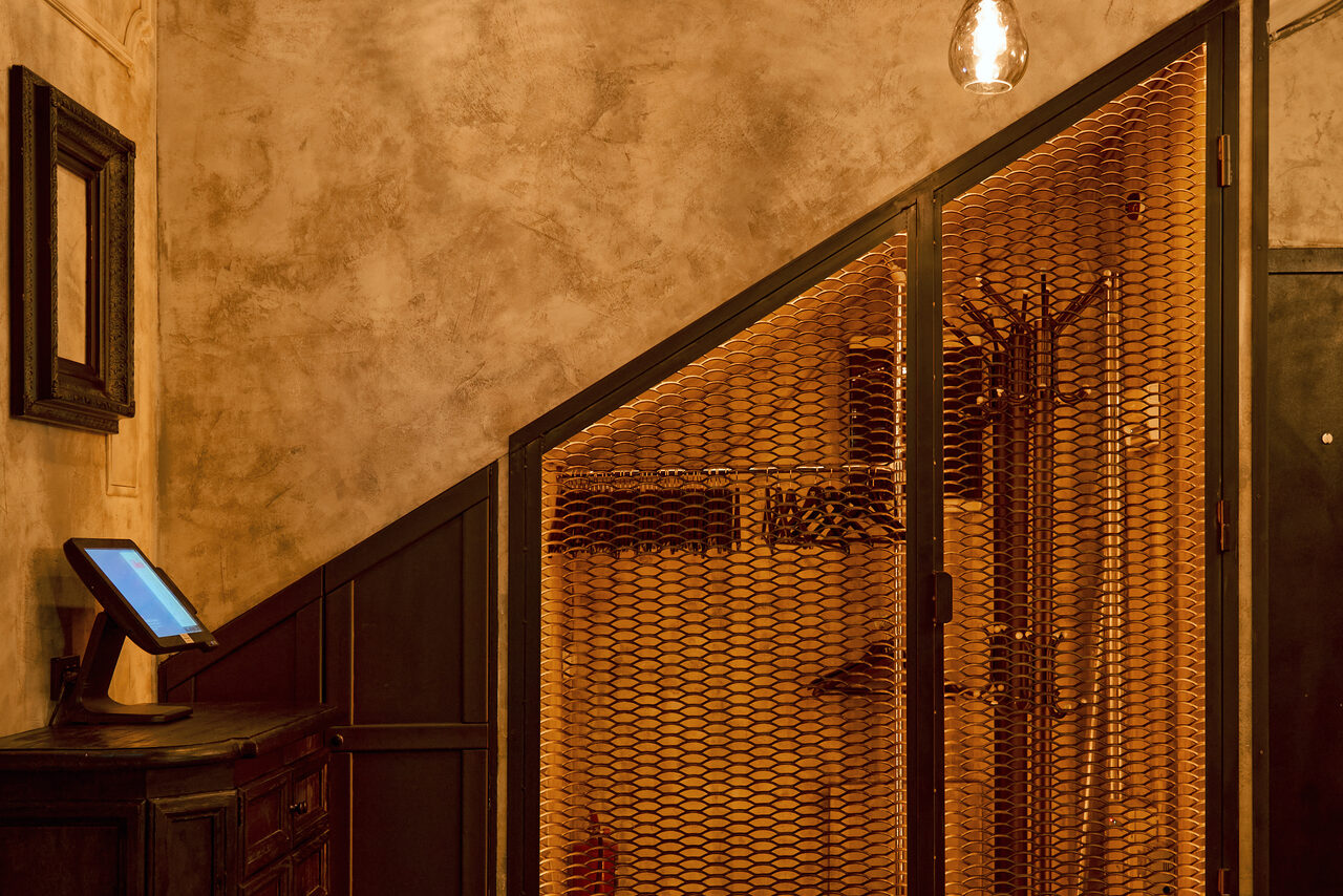 Арендовать Tangiers Lounge(Танжер Лаундж) . Коридор с индустриальными элементами, бежевые стены с текстурой, металлическая сетчатая перегородка, деревянная мебель с монитором, настенный крючок, подвесная лампа с теплым светом, черные и коричневые оттенки, строгая геометрия.