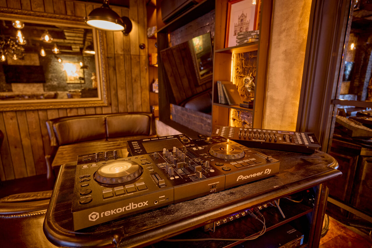 Арендовать Tangiers Lounge(Танжер Лаундж) . Музыкальное пространство с DJ-оборудованием на переднем плане, включая пульт Pioneer Dj и рекордбокс. Индустриальный интерьер с деревянной отделкой, кожаными креслами, полками с книгами и зеркалом. Теплое освещение с подвесными лампами, нейтральные тона.