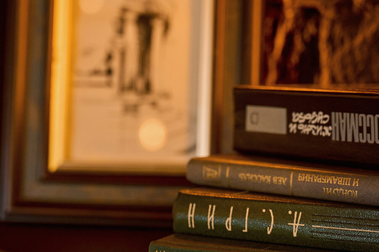 Арендовать Tangiers Lounge(Танжер Лаундж) . Плотно сложенные книги с надписями на корешках на фоне картины в раме с мягким освещением. Цветовая гамма представлена тёплыми оттенками коричневого и зелёного. Видна текстура переплетов книг и деревянной рамы.