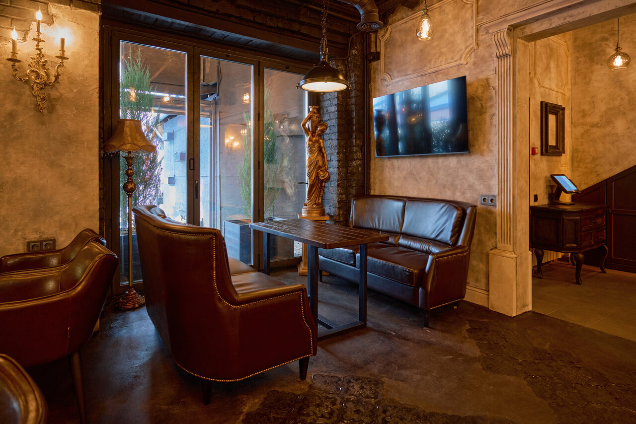 Арендовать Tangiers Lounge(Танжер Лаундж) . Просторный лофт с кожаными диванами и креслами у деревянного стола, большое окно, тёмные подвесные светильники, настенный телевизор, декоративная лампа, светлые стены с текстурой, металлические элементы, высокие потолки, пол тёмного оттенка.