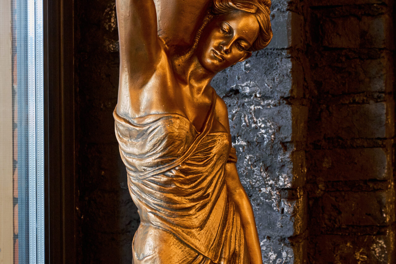 Арендовать Tangiers Lounge(Танжер Лаундж) . Золотистая скульптура женщины в драпировке на фоне черной кирпичной стены у окна. Скульптура изображает женщину с поднятой рукой. Освещение мягкое, оттеняет детали скульптуры и текстуру кирпича.