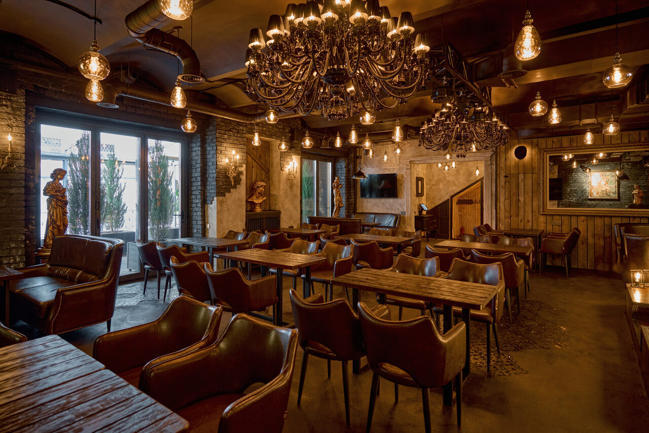 Арендовать Tangiers Lounge(Танжер Лаундж) . Пространство в стиле лофт с деревянными стенами и полом. Столы и кожаные кресла коричневого цвета. Большие люстры и лампы в индустриальном стиле. Декорировано статуями и картинами. Есть окна с плотными шторами. Освещение тёплое.