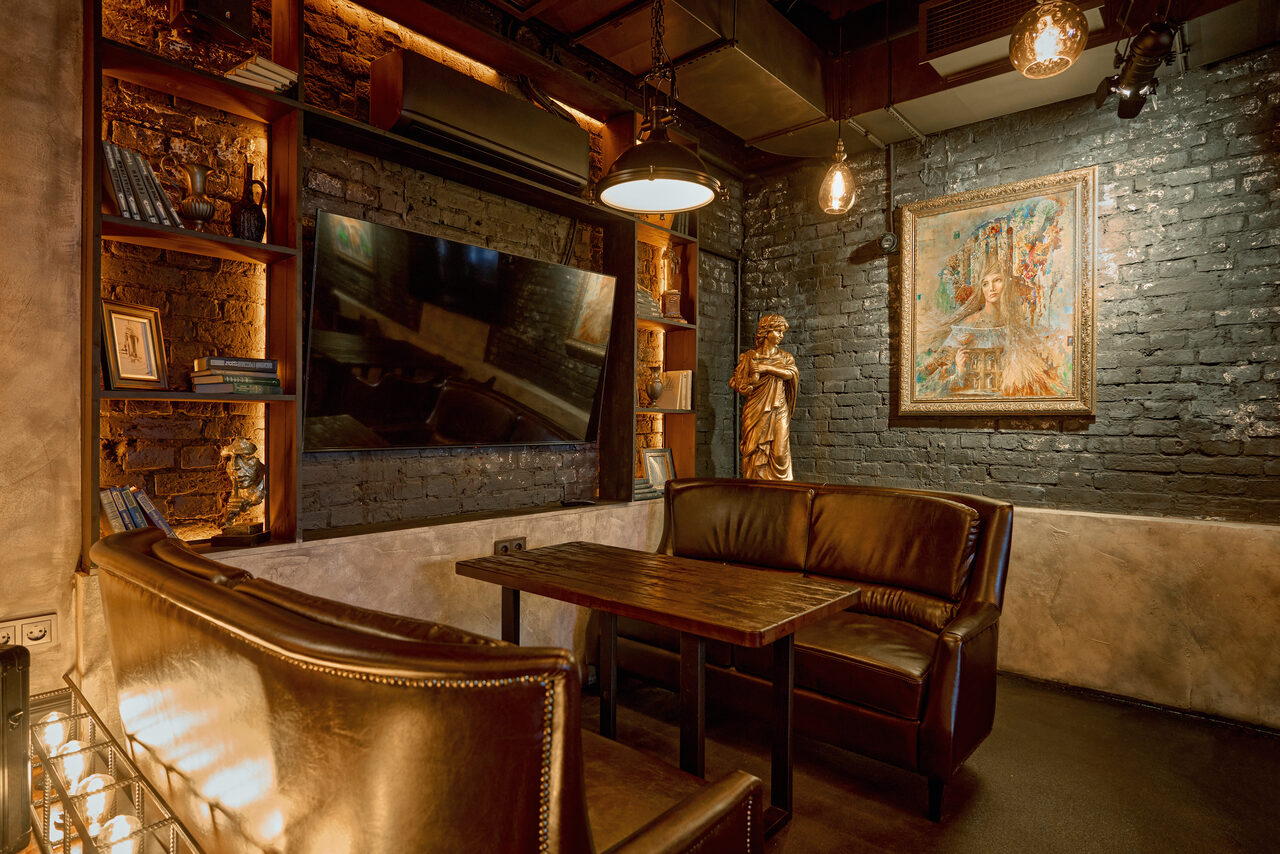 Арендовать Tangiers Lounge(Танжер Лаундж) . Темное уютное помещение с кирпичными стенами. Кожаные диваны у деревянного стола. Картина и статуя в интерьере. Полки с книгами и декором. Подвесные светильники создают теплое освещение. Черный пол, стены частично серые.