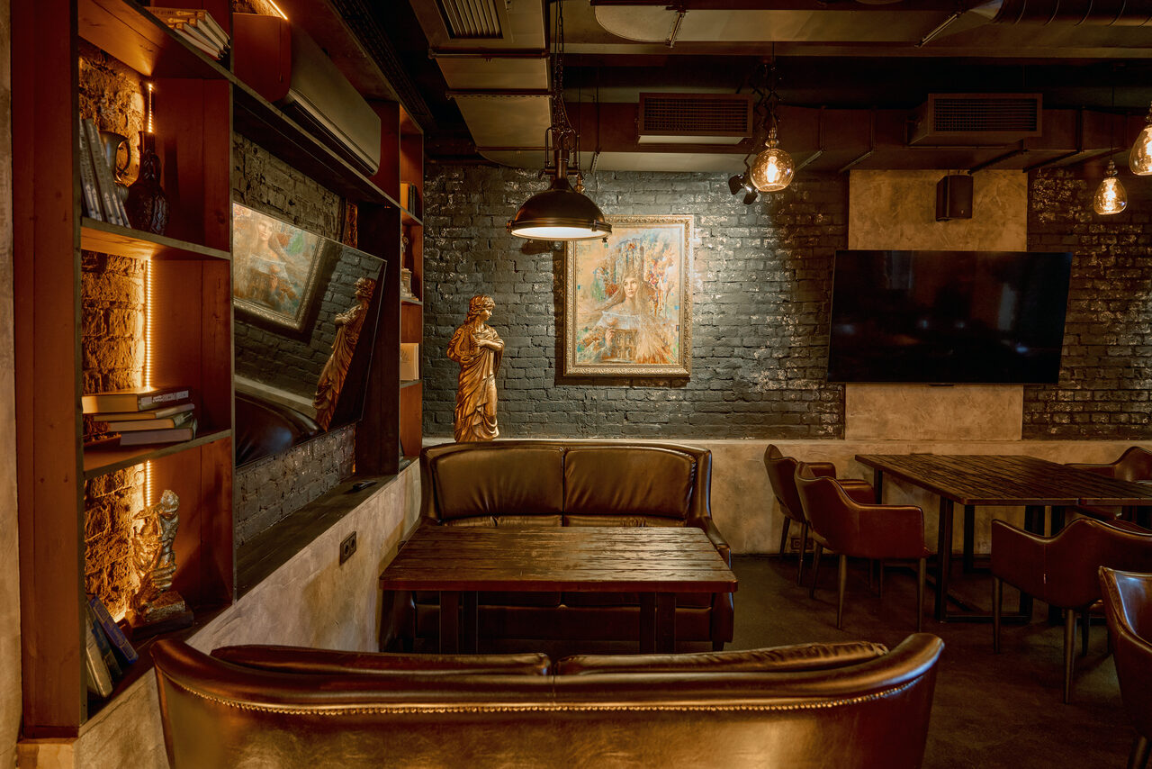 Арендовать Tangiers Lounge(Танжер Лаундж) . Лофт-пространство с темными кирпичными стенами, столами и кожаными креслами. Открытая планировка с деревянными элементами, зеркалом, картинами и статуями. Потолочные светильники создают теплое освещение, общий цветовой тон — коричневый и черный.