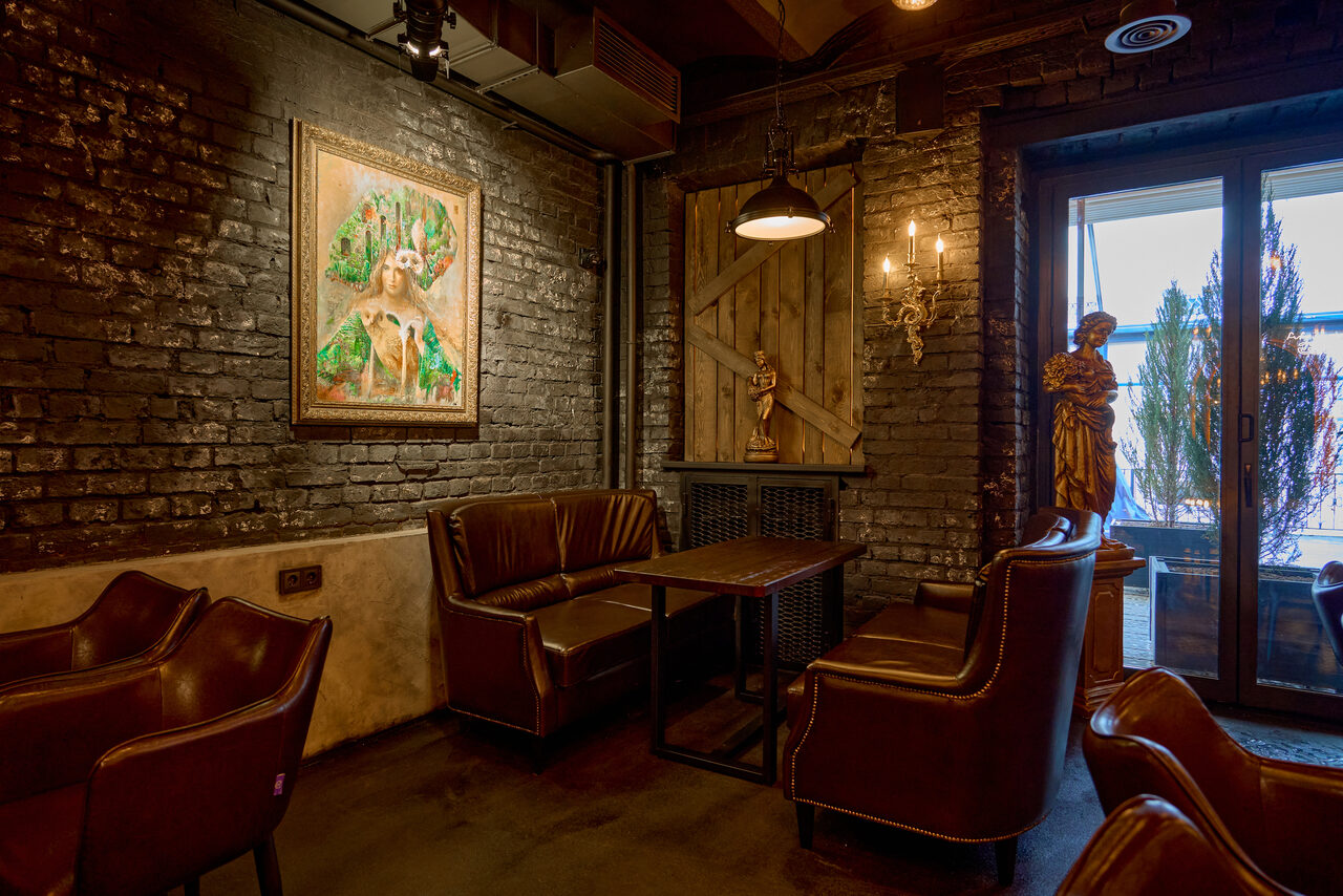 Арендовать Tangiers Lounge(Танжер Лаундж) . Кафе с кирпичными стенами, деревянной мебелью и кожаными креслами. На стене картина и декоративные элементы. Потолочные и настенные светильники. Большое окно ведет на улицу, на подоконнике растения. Тёмные и теплые цвета, уютная атмосфера.