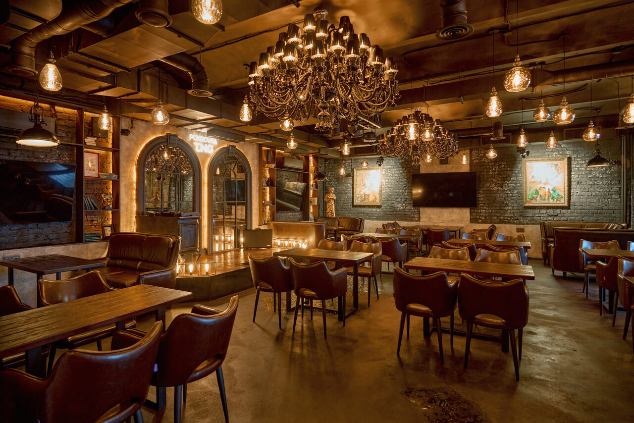 Арендовать Tangiers Lounge(Танжер Лаундж) . Пространство с обеденными столами и кожаными стульями, две большие люстры, множество лампочек накаливания, темные кирпичные стены с картинами, телевизор на стене, барная стойка с арочными зеркалами, свечи на барной стойке, потолок с видимыми трубами.