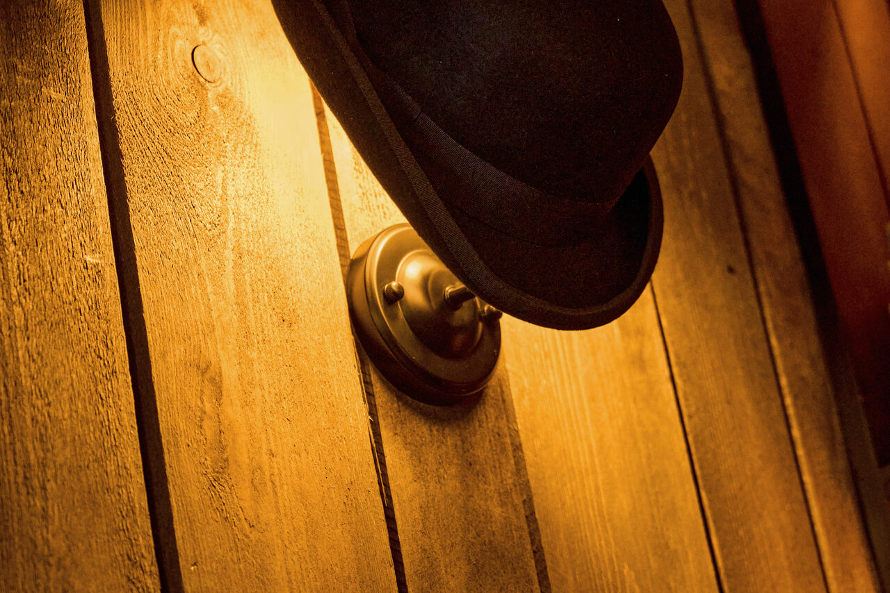 Арендовать Tangiers Lounge(Танжер Лаундж) . Деревянная стена из светлых вертикальных досок, металлический настенный светильник с мягким желтым освещением, черная шляпа на светильнике.