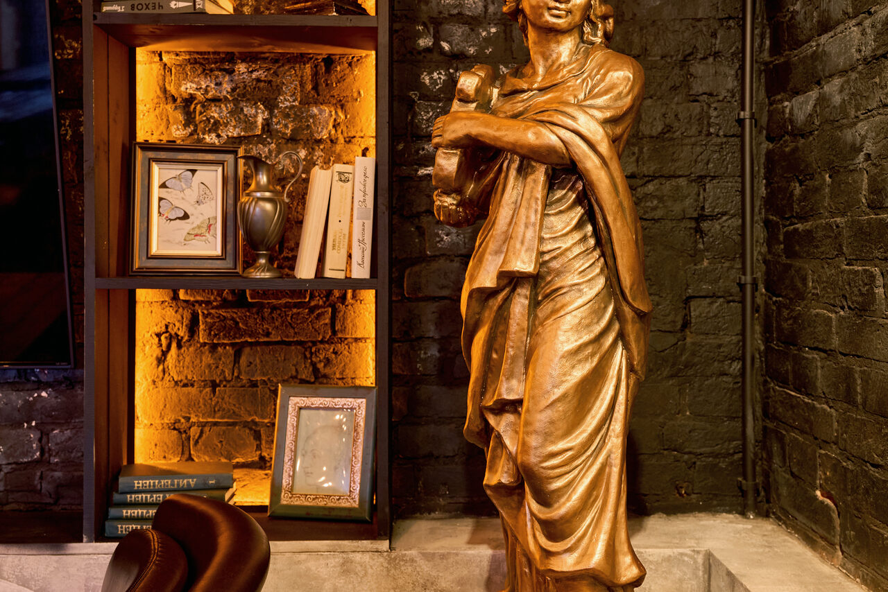 Арендовать Tangiers Lounge(Танжер Лаундж) . Бронзовая статуя в классическом стиле на фоне черной кирпичной стены. Слева встроенный стеллаж с декоративной подсветкой, книгами, рамками и вазой. Стеллаж частично выложен кирпичом. Пол бетонный, цвета теплые, коричневые и черные.