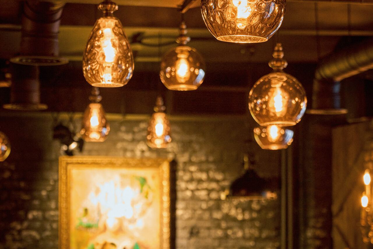 Арендовать Tangiers Lounge(Танжер Лаундж) . Потолочные светильники с теплыми лампами накаливания, стеклянные абажуры, кирпичная стена темного оттенка, картина в раме, металлические вентиляционные трубы, полумрак, индустриальный стиль.