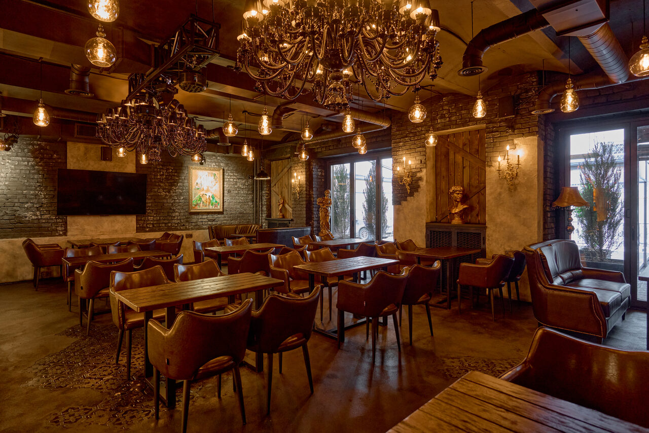 Арендовать Tangiers Lounge(Танжер Лаундж) . Ресторанное пространство с кирпичными стенами, деревянной мебелью, кожаными стульями, круглыми столами. Подвесные лампы и люстры обеспечивают теплое освещение. Присутствуют картины на стенах и статуи. Металлические трубы и вытяжки на потолке.