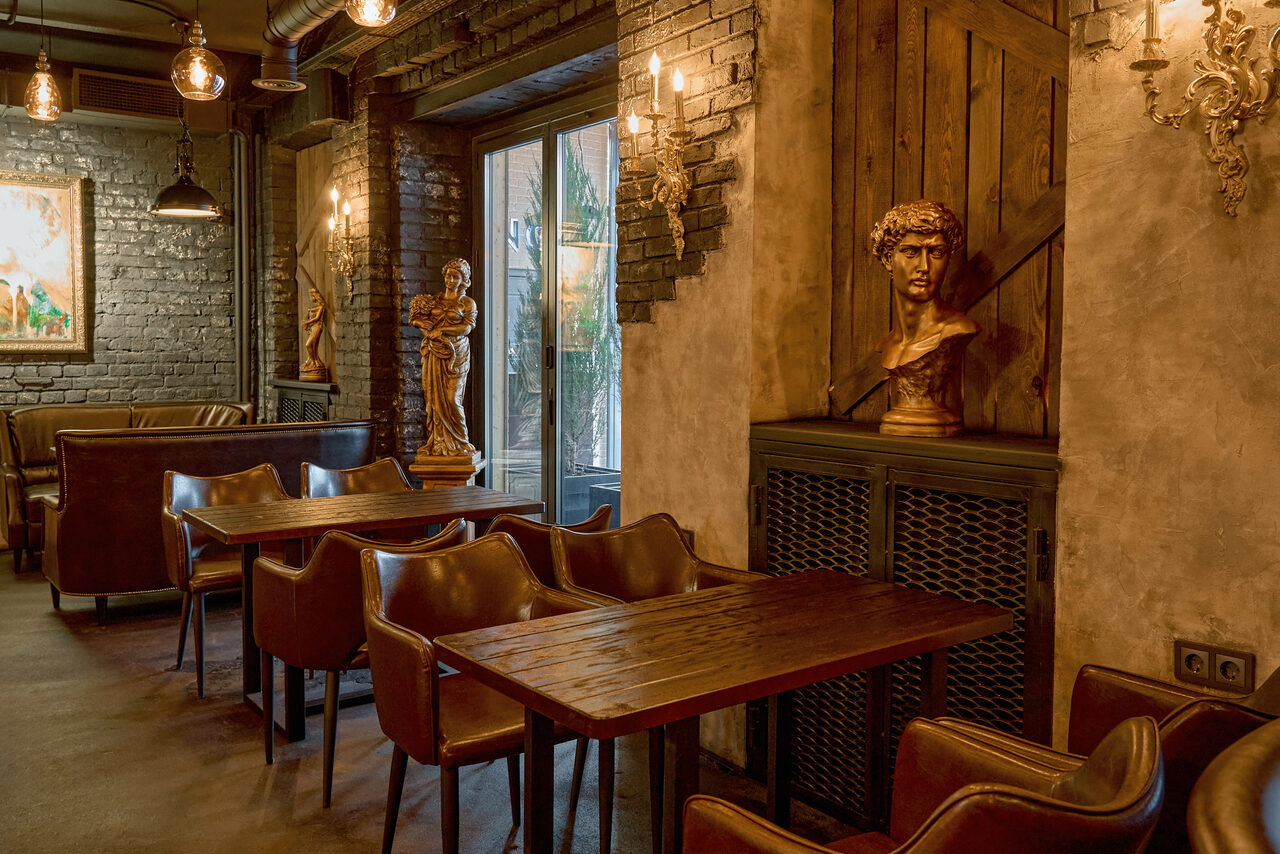 Арендовать Tangiers Lounge(Танжер Лаундж) . Лофт-пространство с кирпичными стенами, деревянными столами и кожаными стульями. Настенные светильники и лампы на потолке обеспечивают теплое освещение. Декор включает статуи и бюсты, большие окна, картина на стене. Доминируют коричневые и серые тона.