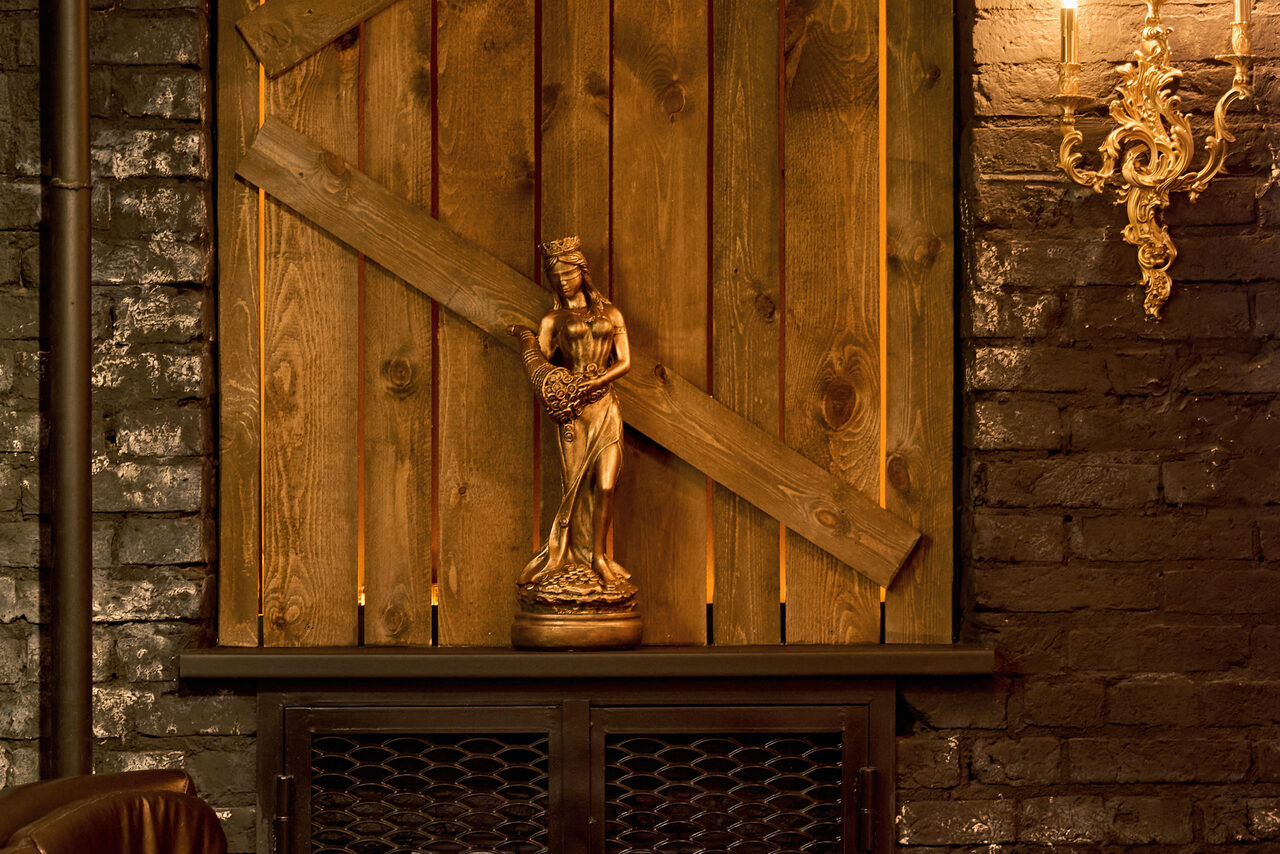 Арендовать Tangiers Lounge(Танжер Лаундж) . Деревянные панели на стене с декоративной статуэткой, расположенной на деревянной полке. Коричневое кирпичное покрытие стен, металлический светильник золотого цвета на стене слева. Теплое освещение.