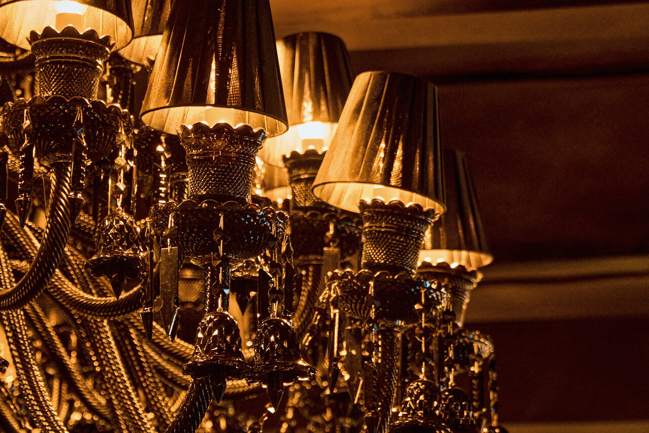 Арендовать Tangiers Lounge(Танжер Лаундж) . Классическая люстра с несколькими лампами в металлических абажурах, украшенная декоративными элементами и цепями. Тёплое освещение, преобладание тёмных и золотых тонов.