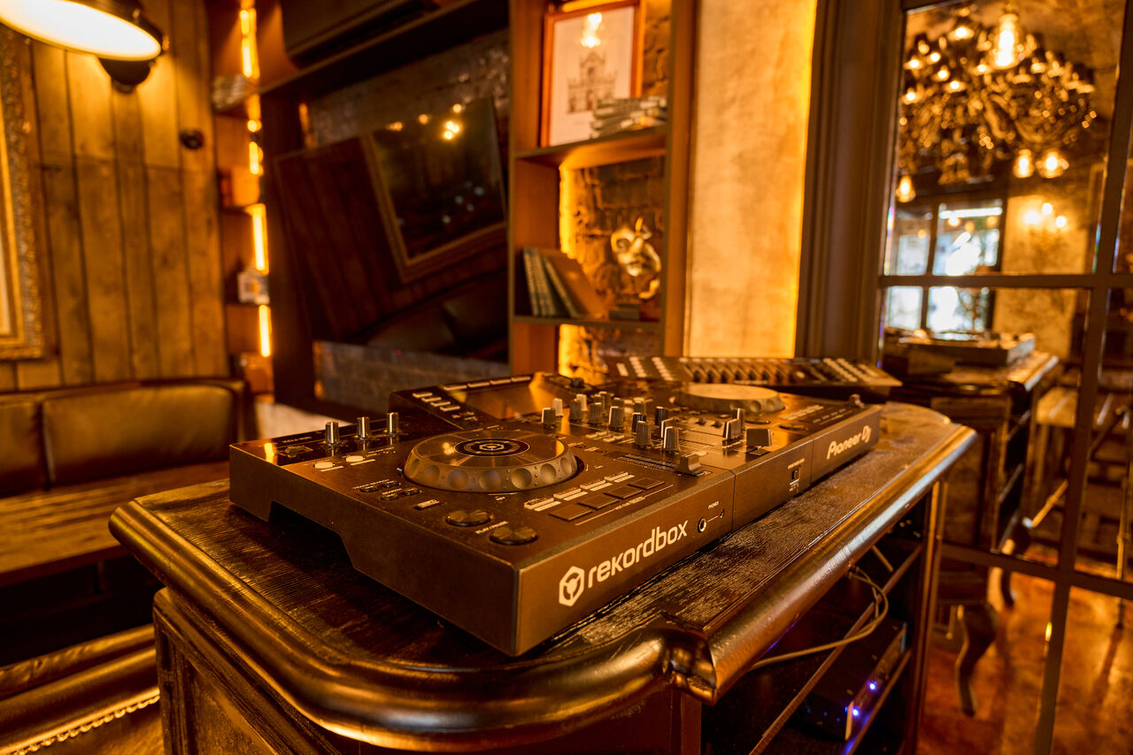 Арендовать Tangiers Lounge(Танжер Лаундж) . DJ-оборудование на стойке из тёмного дерева, кожаный диван, деревянные панели на стене. Тёплое освещение с подвесными лампами, книжные полки, зеркала. Декоративные элементы и картины, отделка под кирпич. Тёплые коричневые и золотистые тона.