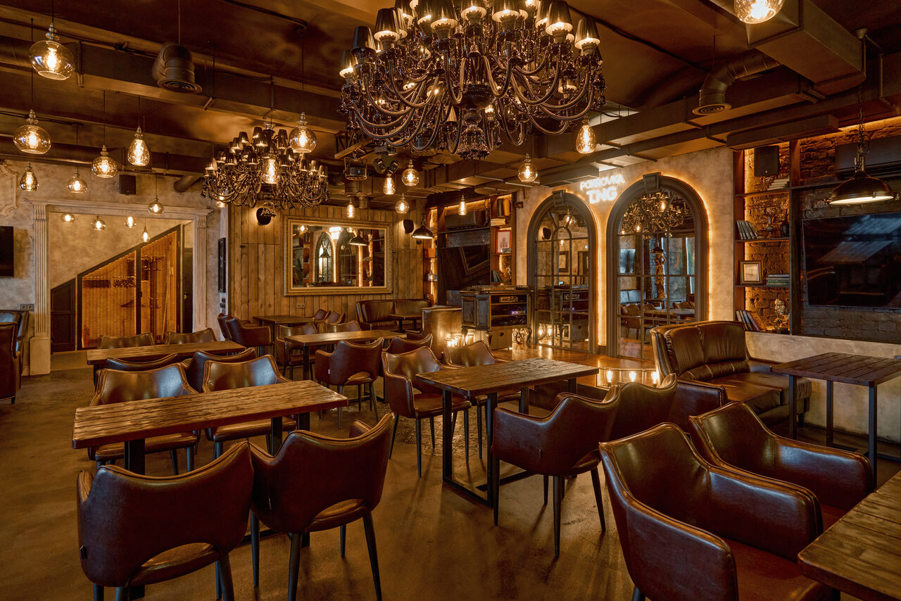 Арендовать Tangiers Lounge(Танжер Лаундж) . Барное пространство с деревянными столами и кожаными стульями коричневого цвета. Потолочные люстры и лампы в индустриальном стиле. Стены отделаны древесиной и камнем, есть зеркала и арочные дверные проёмы. Теплое освещение создает уютную атмосферу.