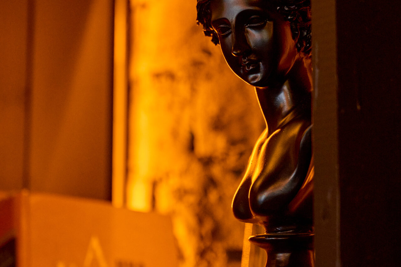 Арендовать Tangiers Lounge(Танжер Лаундж) . Скульптура женского бюста в тёмном цвете на фоне мягкого оранжевого освещения и нечетких текстур. Боковая часть стены разделяет композицию, виден фрагмент картонной коробки.