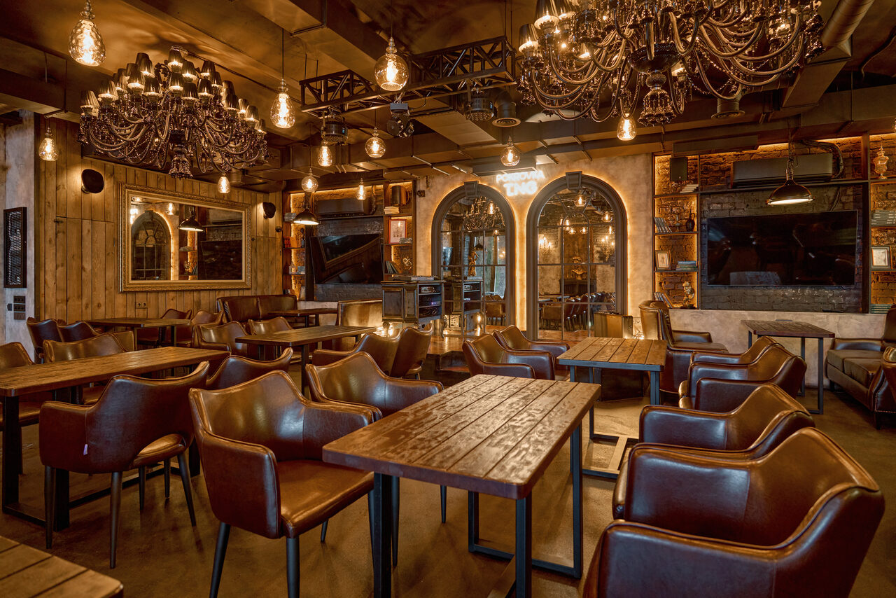 Арендовать Tangiers Lounge(Танжер Лаундж) . Ресторанное пространство с деревянными столами и коричневыми креслами. Подвесные светильники и большие люстры создают теплое освещение. Индустриальный стиль с элементами кирпичной кладки и деревянной обшивки. Арочные двери, зеркала, декоративные полки.