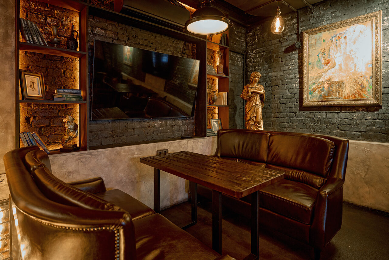 Арендовать Tangiers Lounge(Танжер Лаундж) . Темная комната с кирпичными стенами, кожаные диваны коричневого цвета, деревянный стол, настенный телевизор, картина в раме, статуя, открытые полки с декором и книгами, подвесные лампы теплого света.
