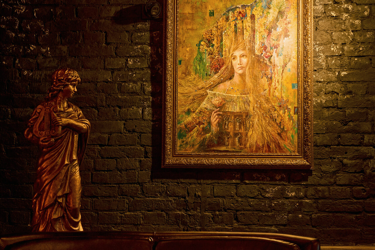 Арендовать Tangiers Lounge(Танжер Лаундж) . Темное пространство с кирпичной стеной, украшенной картиной в золотой раме, изображающей женщину с длинными волосами. Слева стоит скульптура, также покрытая золотым оттенком. Освещение теплое, рассеянное.